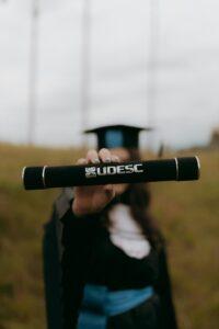Graduate in cap and gown holding diploma outdoors, focus on diploma.
